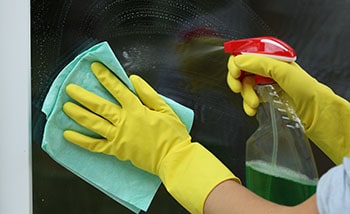 A woman cleans the window by spraying a glass cleaning solution and wiping it with a microfiber cloth.